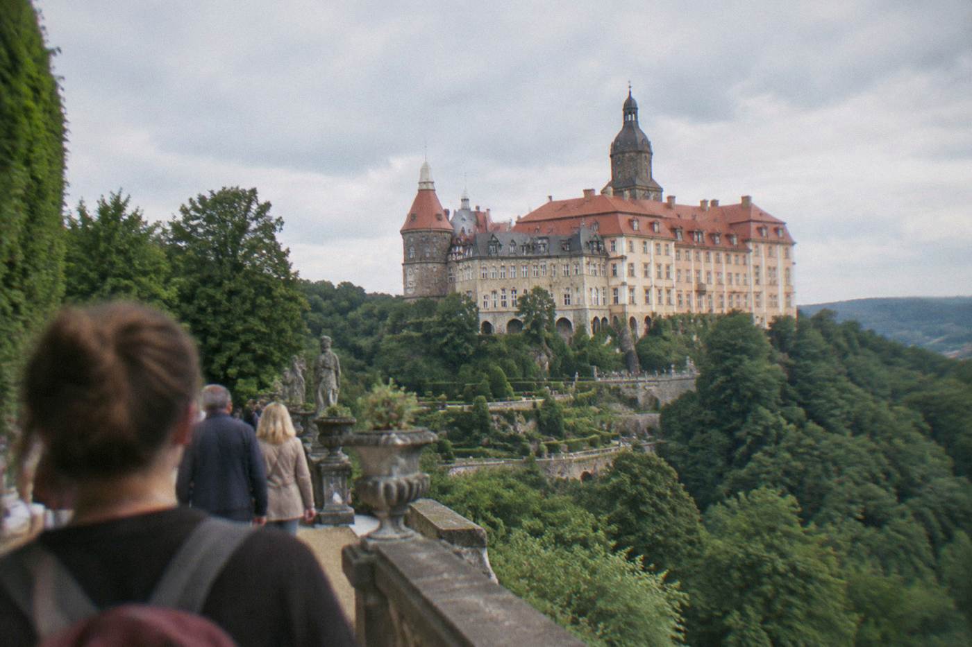 Large cream-colored Ksiaz Castle with red tiled roofs seen from its terraced gardens on a forested hillside
