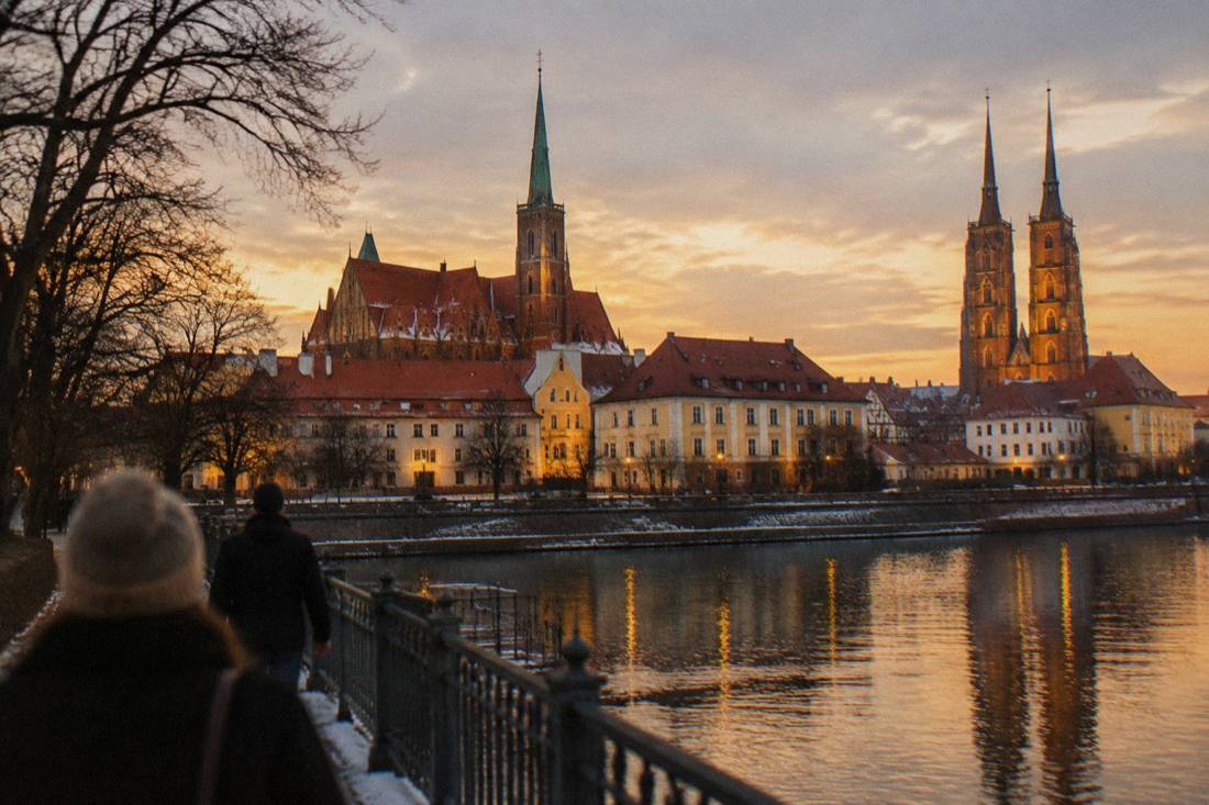 Illuminated historic townhouses and a cathedral tower seen across a river on a winter evening