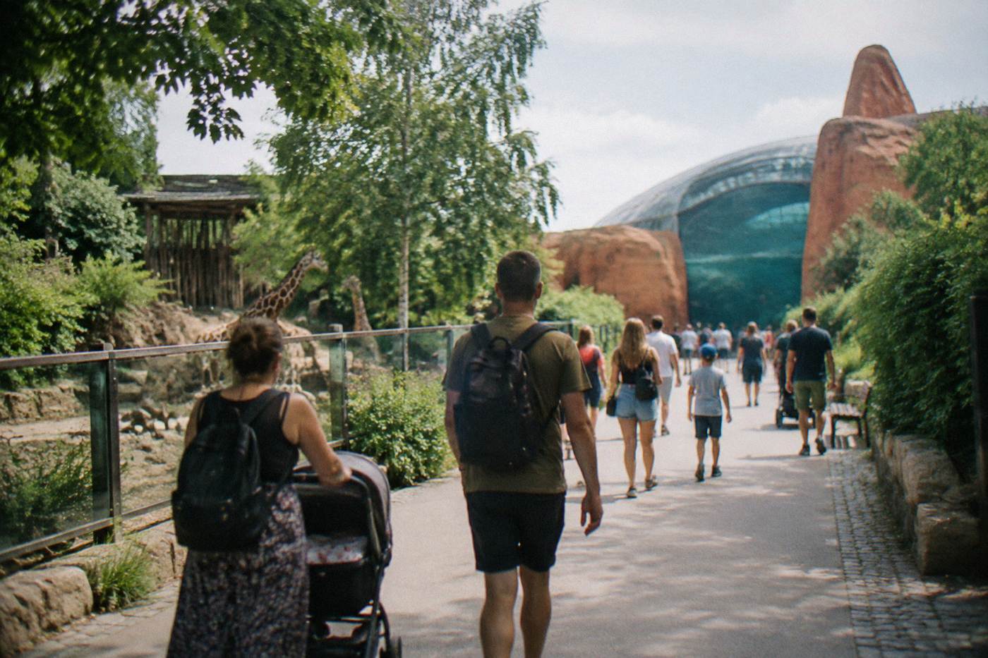 Visitors walking a wide paved path past leafy enclosures on a sunny day at a large city zoo