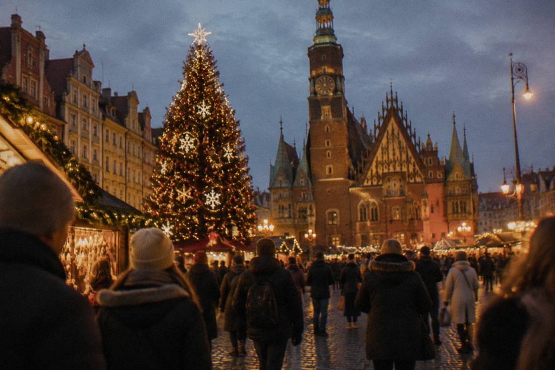 A winter market square with a decorated Christmas tree and crowds in coats at nightfall