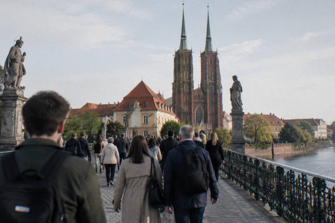 A crowd of pedestrians crossing the stone Tumski Bridge toward the cathedral island on a bright morning
