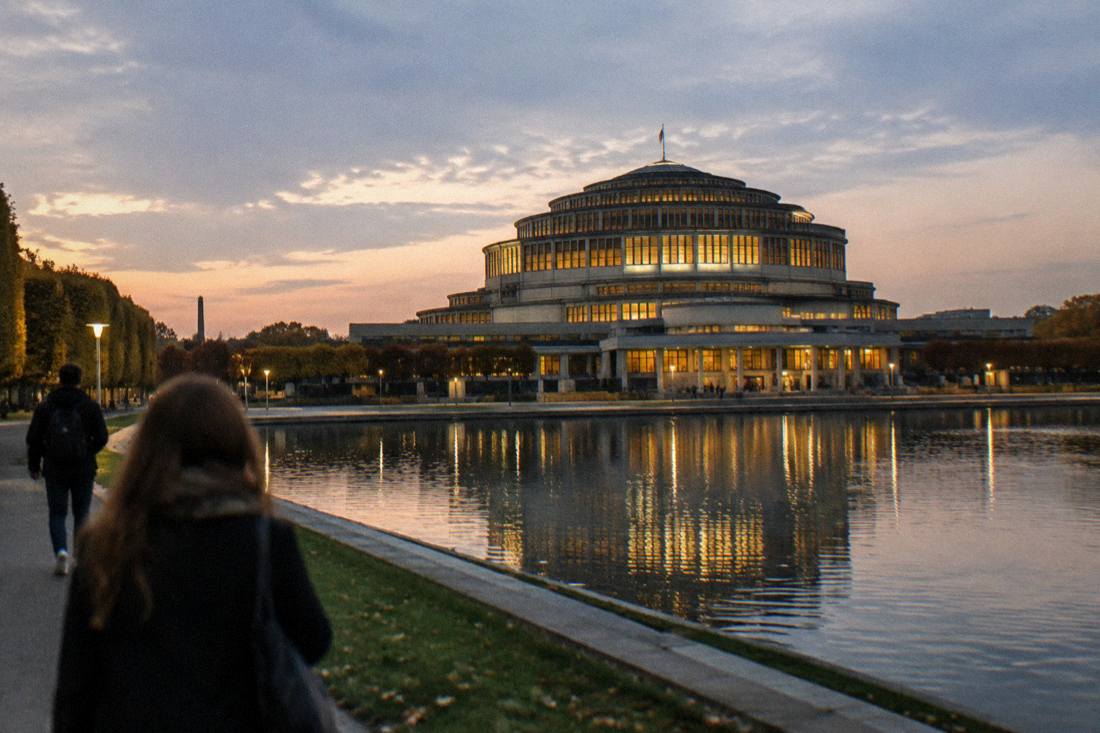 Wide view of a monumental domed hall lit at dusk beside a calm pool of water
