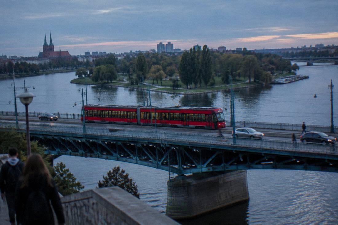 A red tram crossing a steel bridge over a wide river with island parks behind it