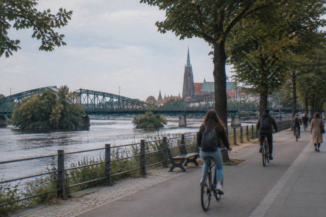 A wide riverside walkway with trees, cyclists and a distant cathedral spire