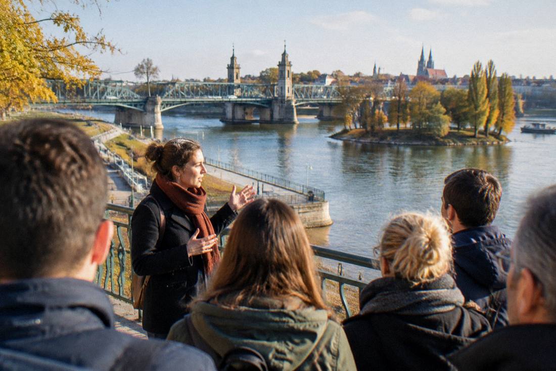 A group of visitors listening to a tour guide beside a river railing with a bridge behind