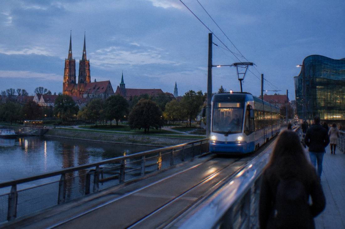 A tram crossing a bridge over the Odra river toward green parkland and old buildings