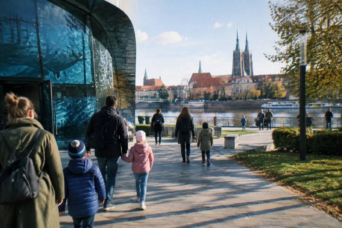Families with children walking out of a modern museum entrance toward a park with the river behind