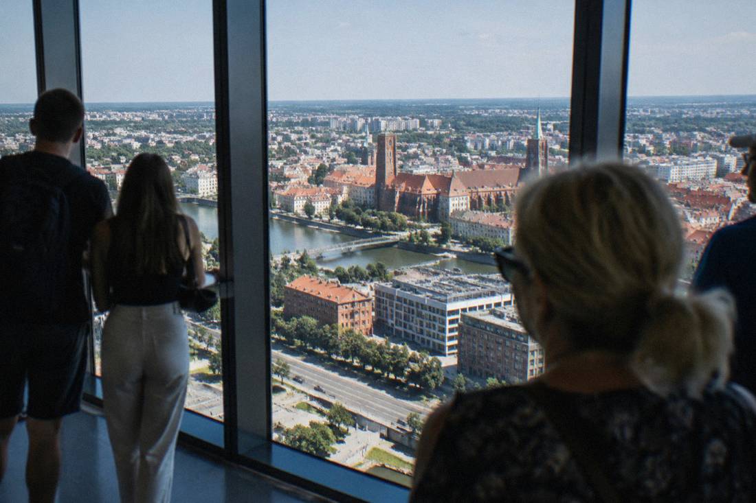 Visitors looking out through tall windows toward the city skyline from a high indoor viewing level