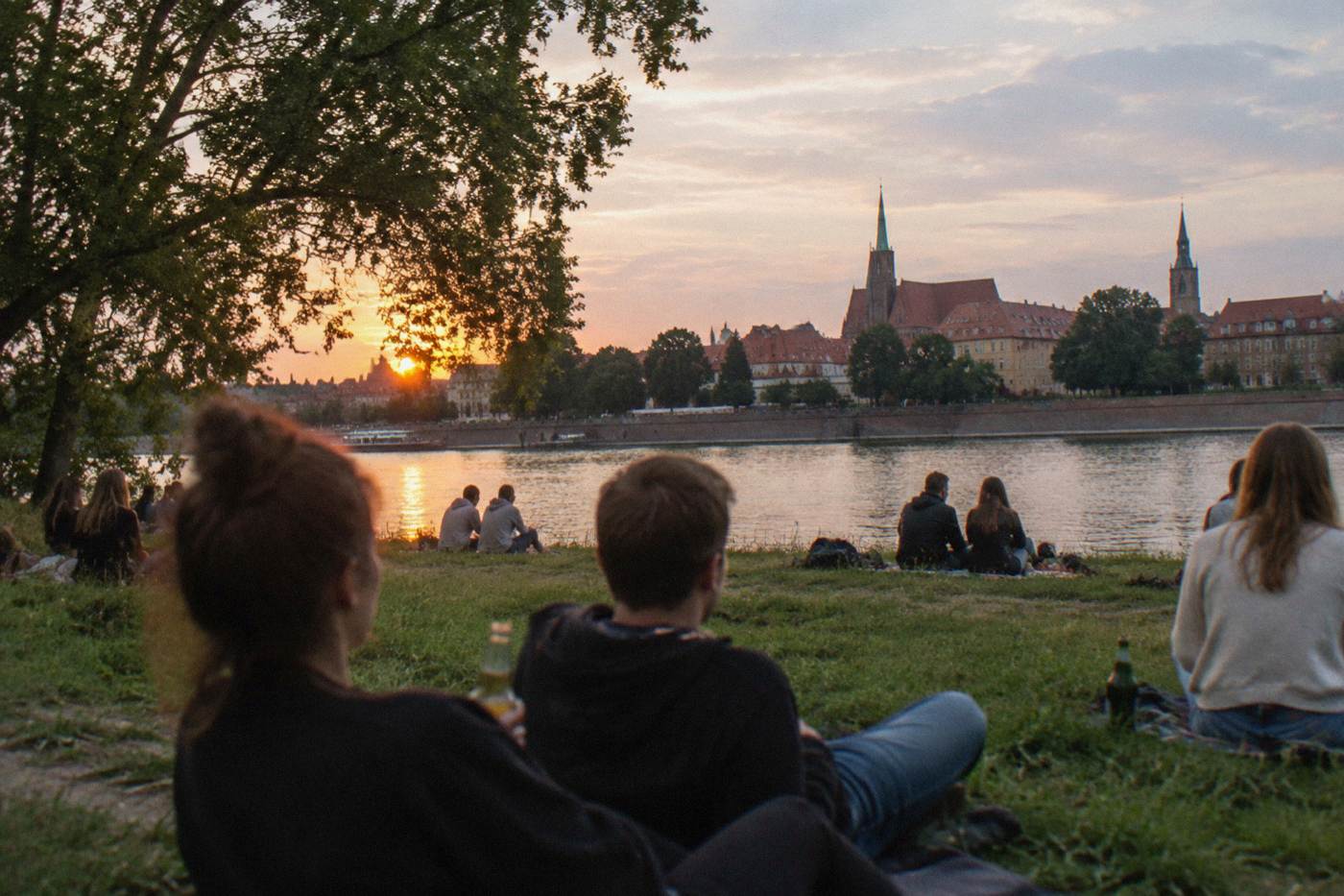 People relaxing with drinks on a grassy river island at sunset, with trees and old city rooftops across the water