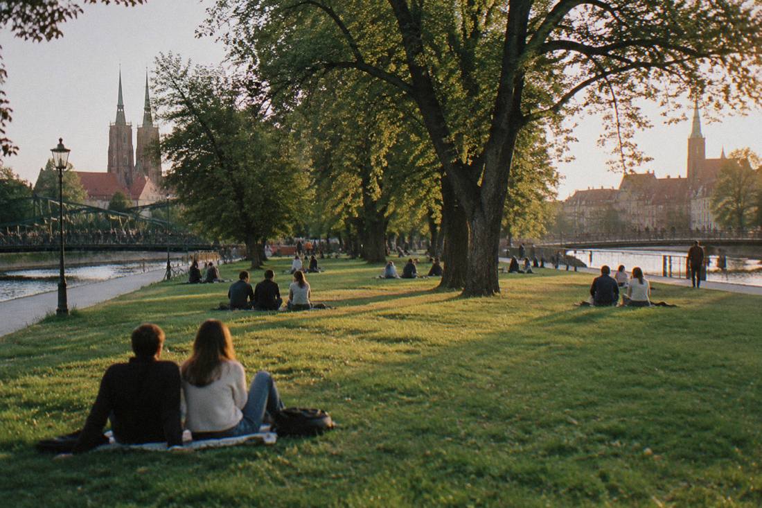 A wide green lawn on a river island with mature trees, footbridges at each end and people sitting on the grass