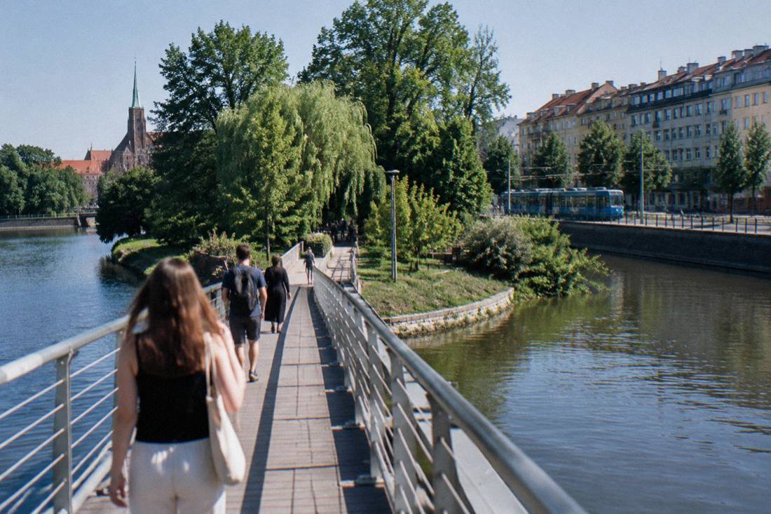 A footbridge over a narrow river channel leading toward a green island, with tram tracks and apartment buildings on the far bank