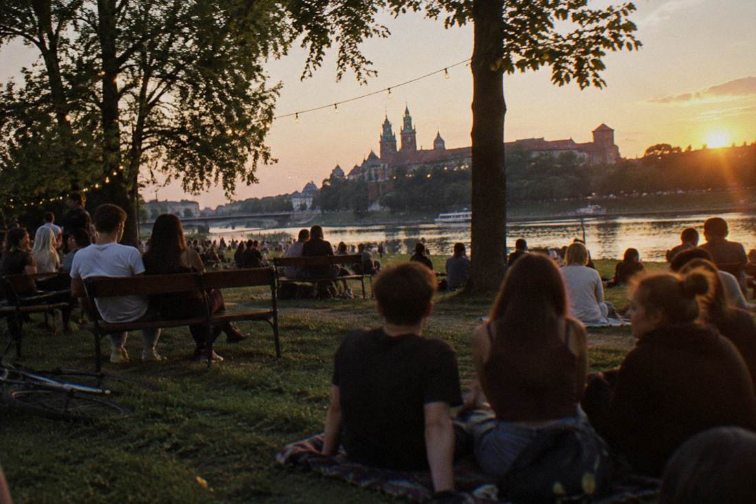 A crowd of young people sitting on benches and grass at dusk on a river island, string lights and trees visible