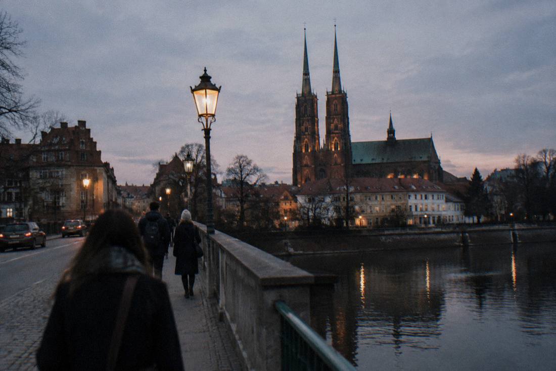 A stone bridge over a river channel at dusk, with gas-style lanterns and the silhouette of a cathedral beyond
