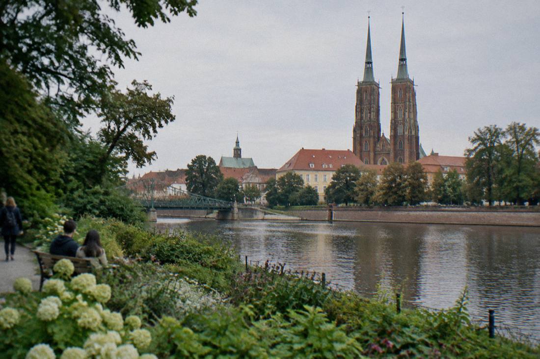 A historic cathedral with twin spires seen across a river from a green park embankment
