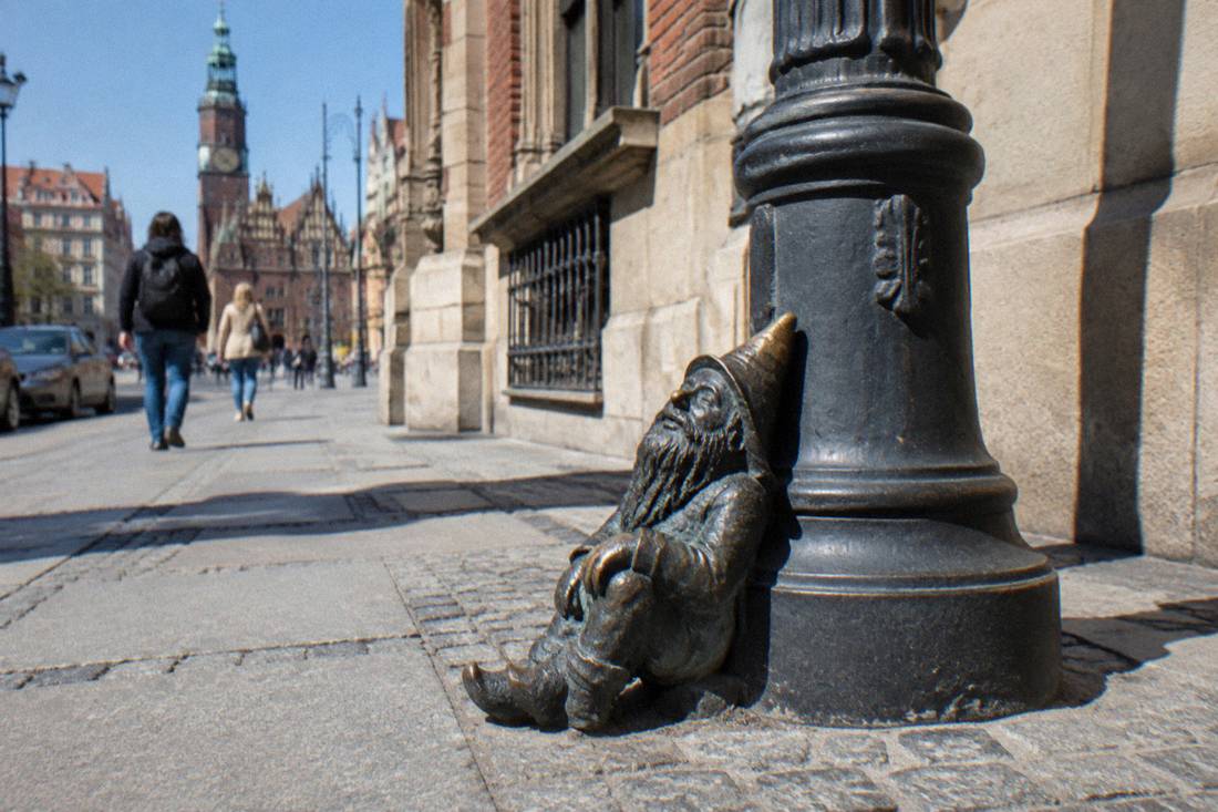 A bronze dwarf figure resting on the base of a lamppost beside a historic building facade