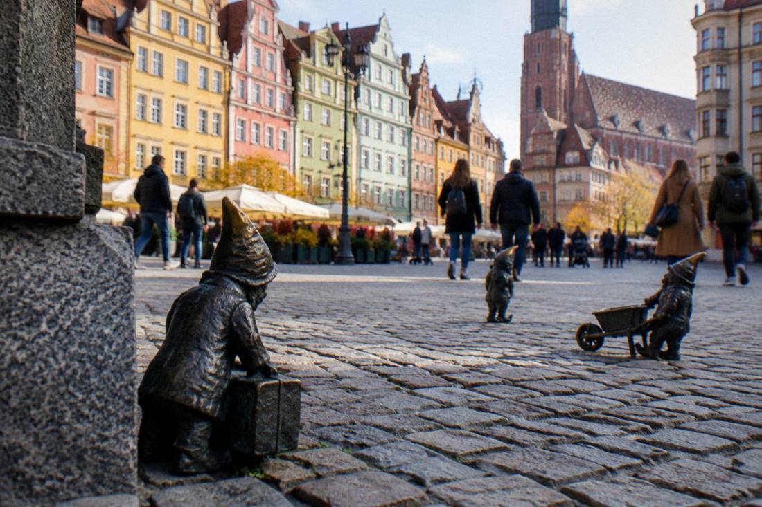 A row of colorful townhouses above a cobbled market square where tiny bronze figures hide