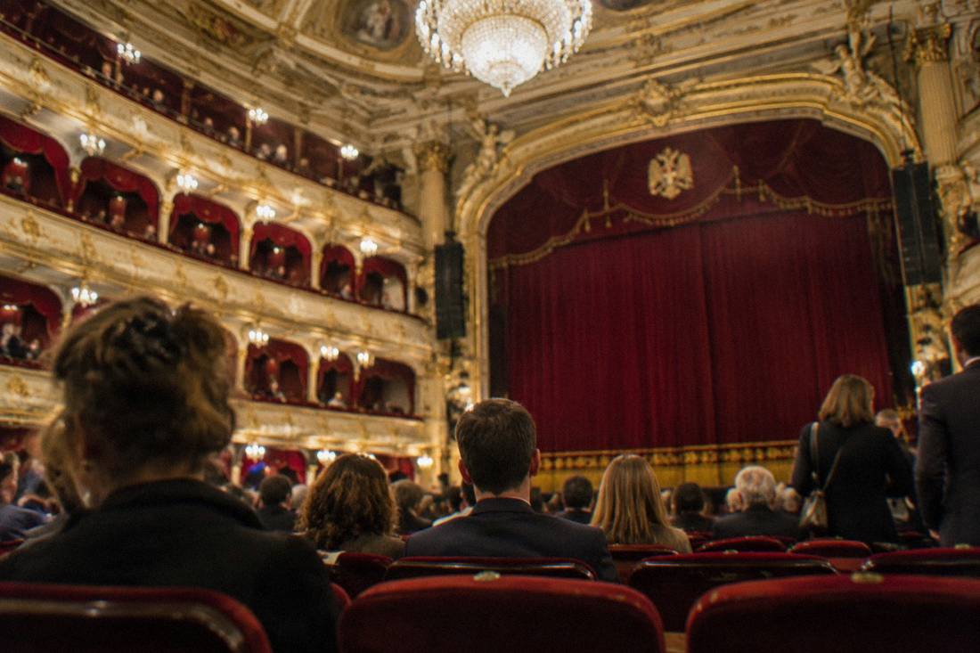 Theater auditorium with rows of red velvet seats, gilded balconies and a glowing chandelier