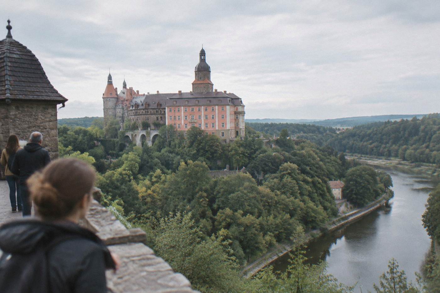 A large Baroque castle on a forested hill above a river bend in Lower Silesia, Poland