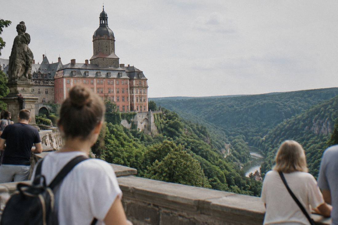 Visitors on a castle terrace overlooking a green forested river valley in summer