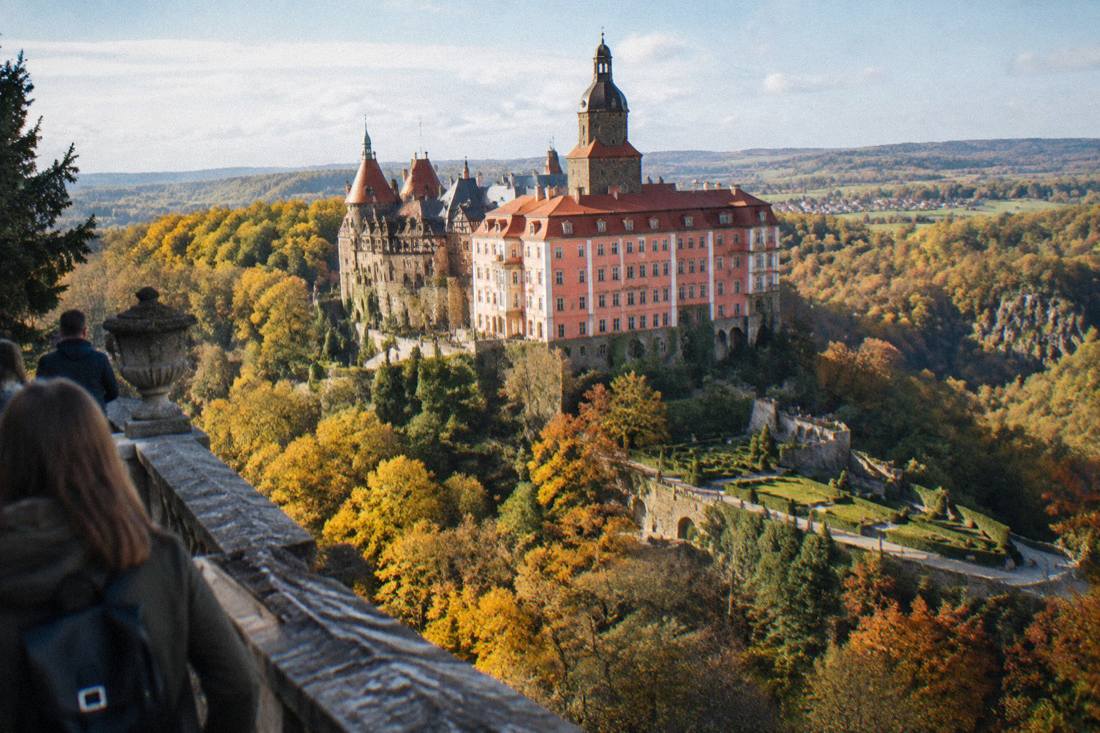 Aerial-style view of a large castle complex with gardens rising above a forested valley in Lower Silesia