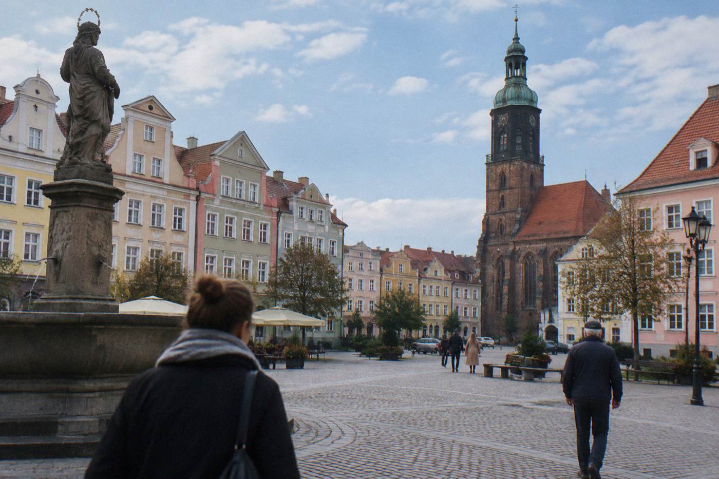 A quiet Polish town square with pastel townhouses and a church tower under a blue sky