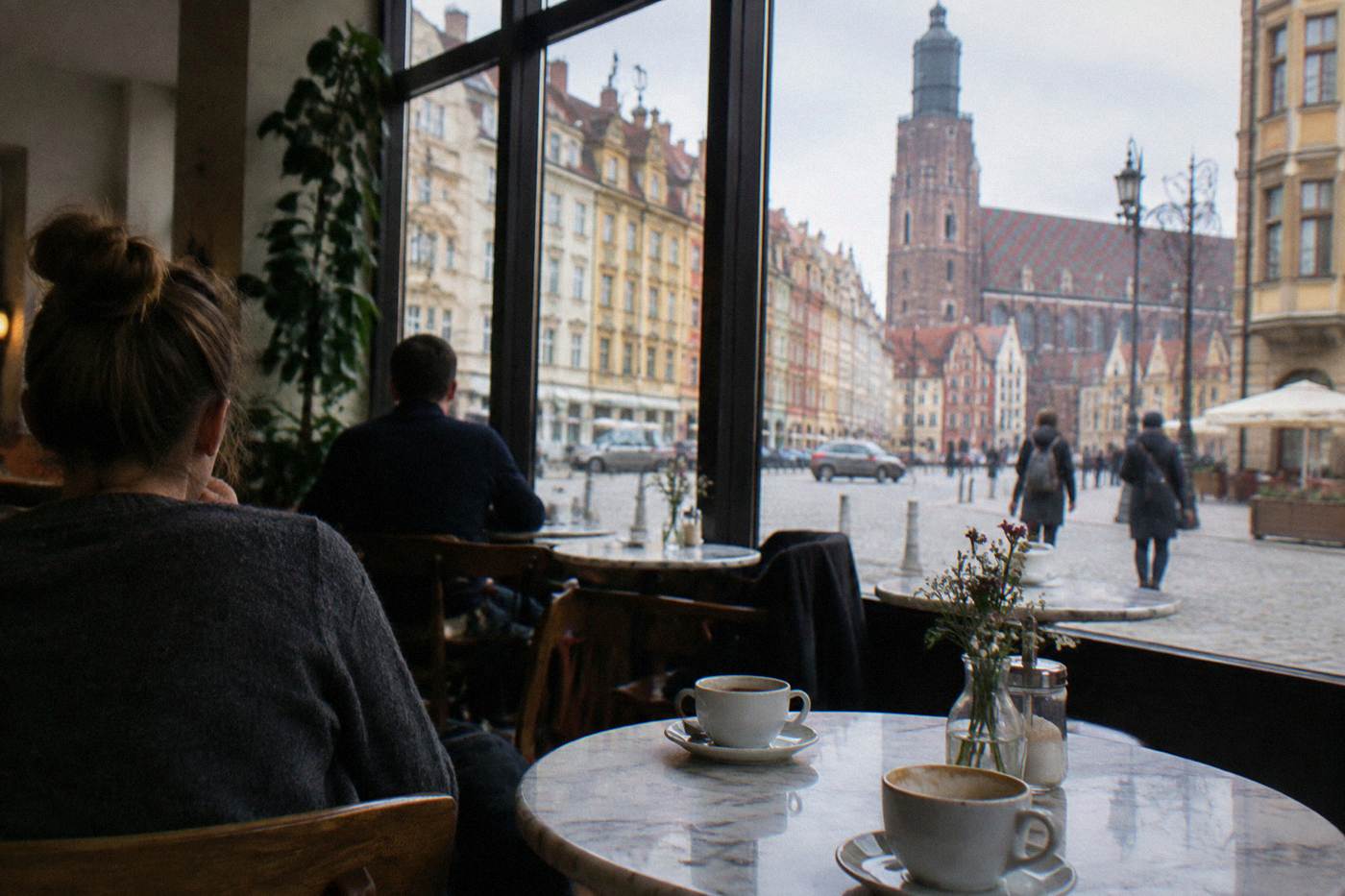 A quiet cafe interior with small marble tables, cups of coffee, and tall windows looking onto a European old town street