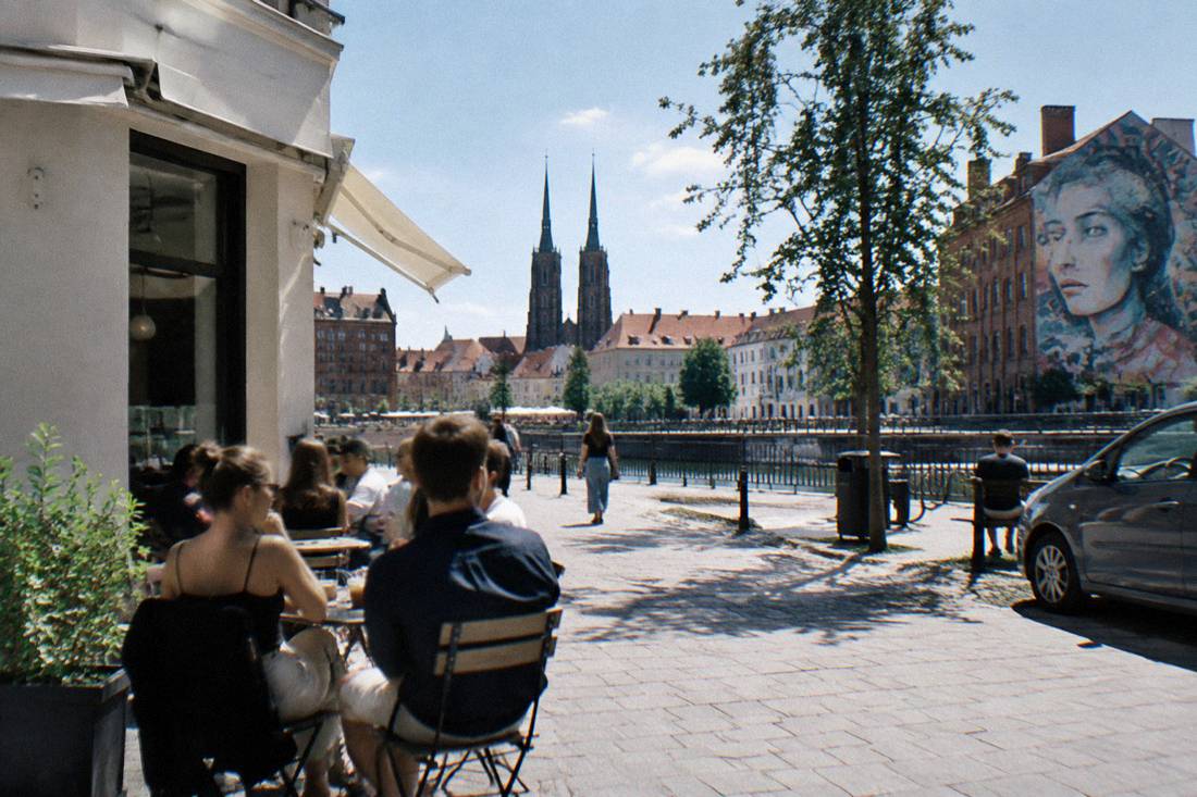 People sitting outside a bright corner cafe in a revitalized city district with murals on nearby walls
