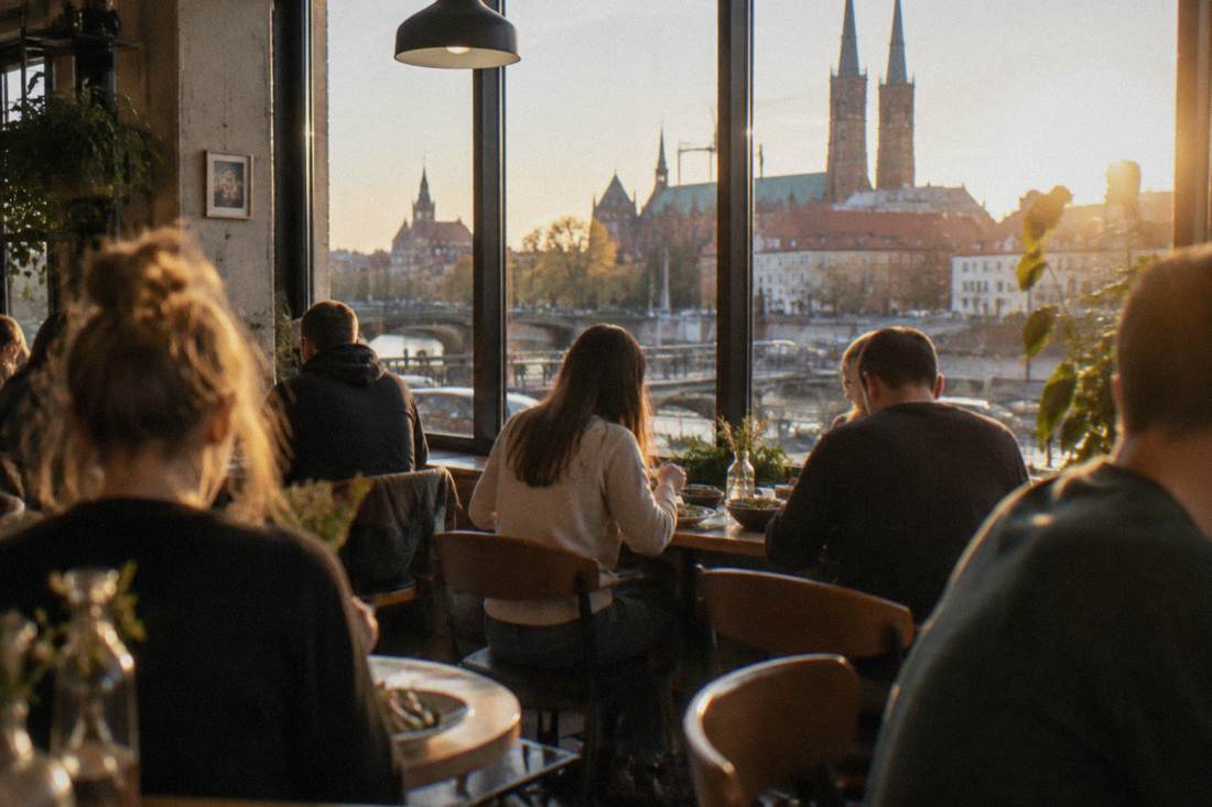 A busy cafe interior with wooden chairs and people eating lunch near a large window