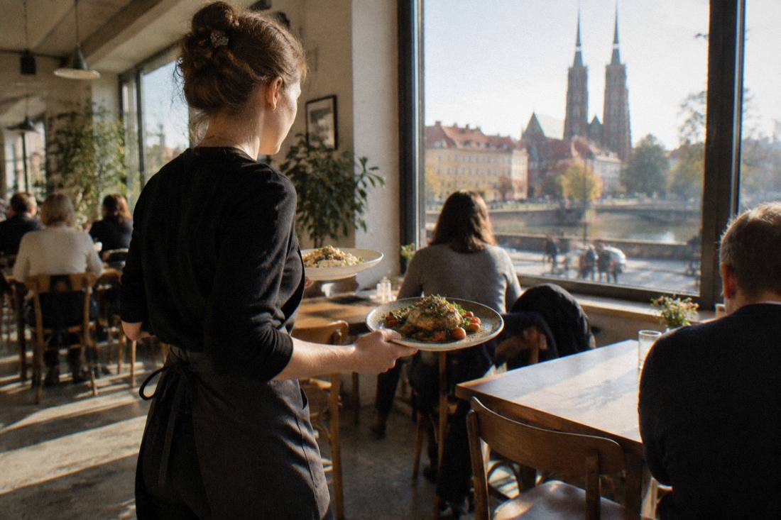 A server carrying two plates of food to a table in a brightly lit bistro