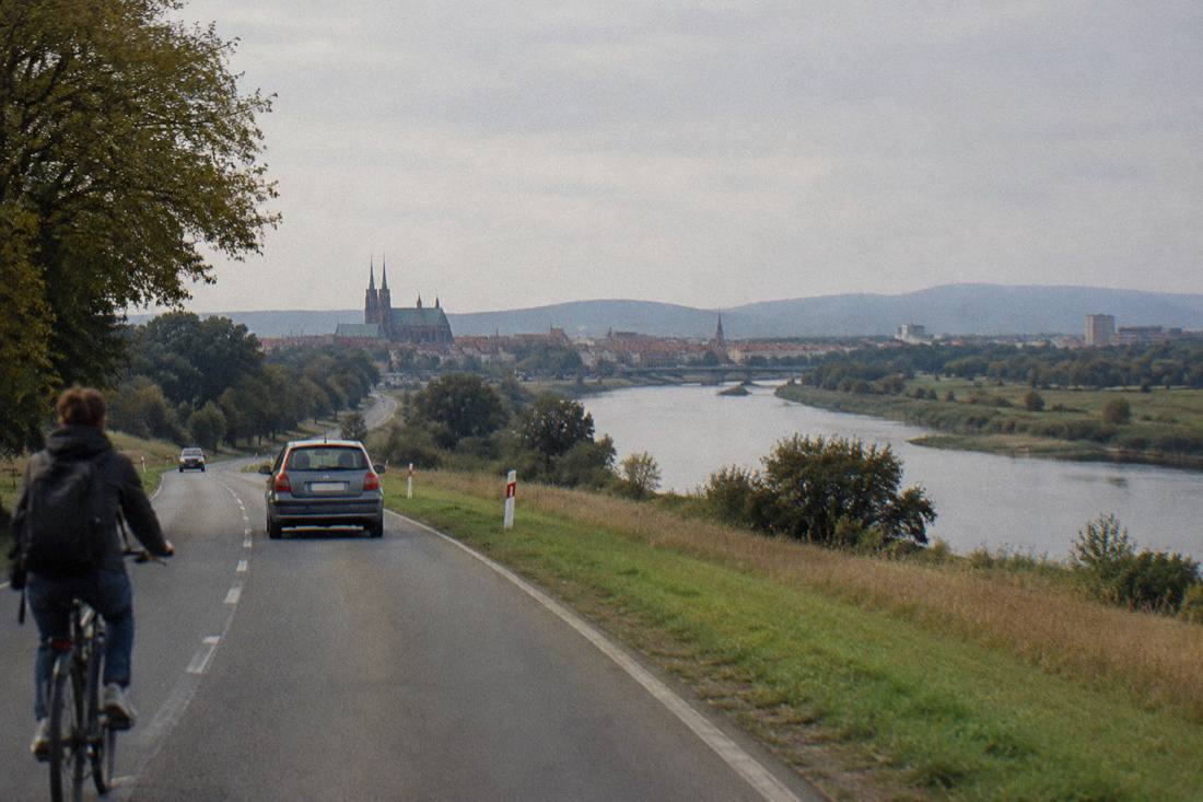 An open road crossing a rural landscape with a border-area river and hills on the horizon