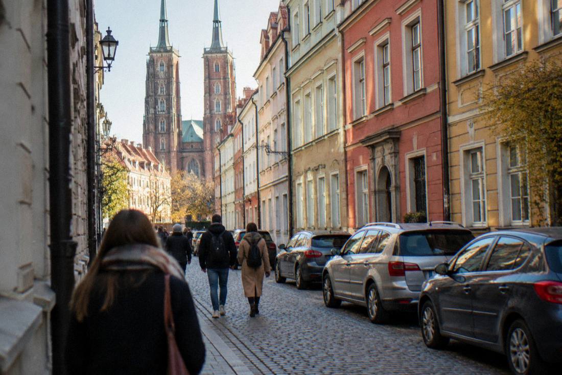 A narrow Wroclaw old town street with pedestrians and a few parked cars between townhouses