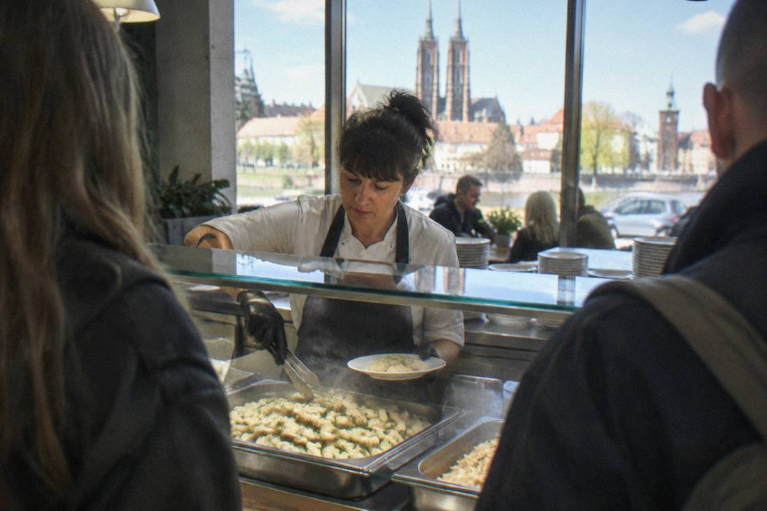 A cafeteria counter worker serving dumplings from a steaming tray behind a glass display
