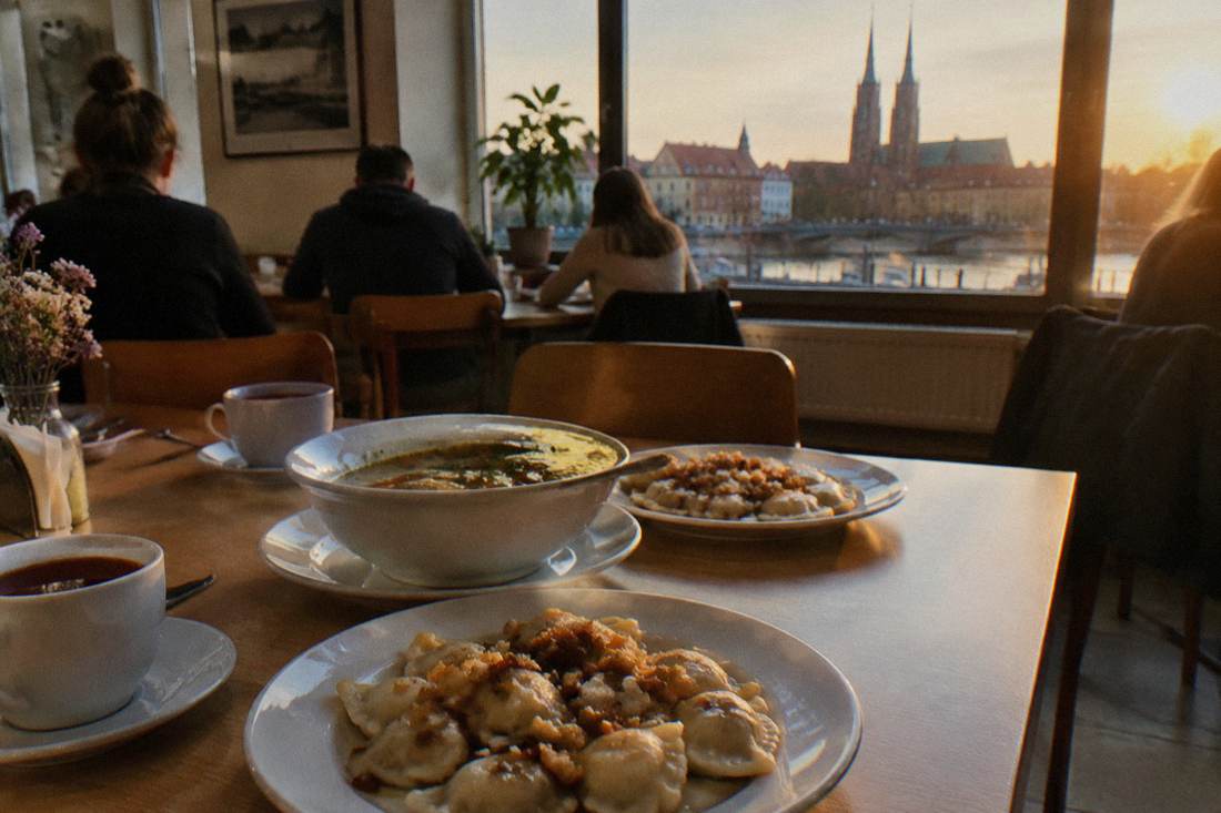 A cafeteria table with plates of Polish dumplings, a bowl of soup and cups of tea in a simple dining room