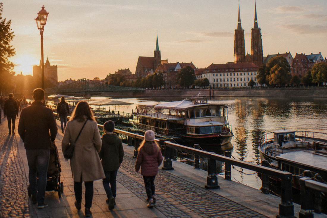 Families strolling on a riverside embankment in Wroclaw with moored boats visible along the bank