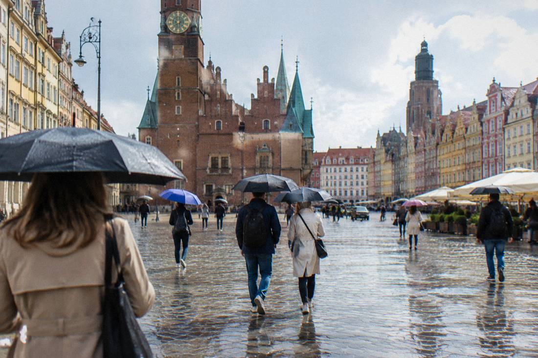 Visitors walking with umbrellas across a European old town square in light rain