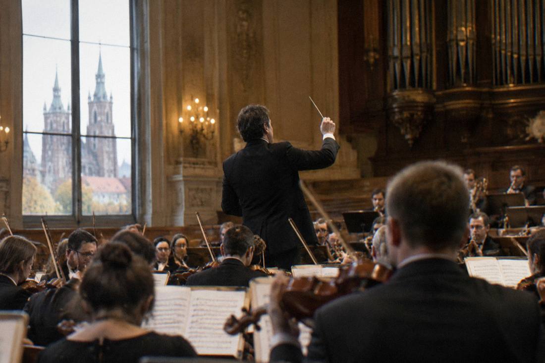 A conductor raising a baton before a full orchestra in a lit concert hall