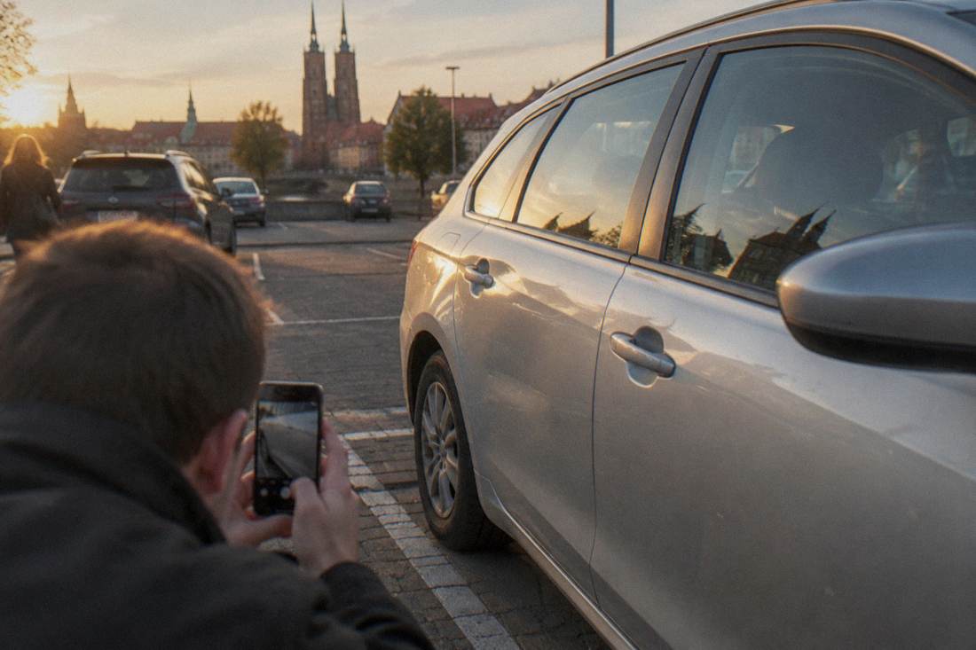 A driver photographing the side panel of a silver rental car in an outdoor parking area.