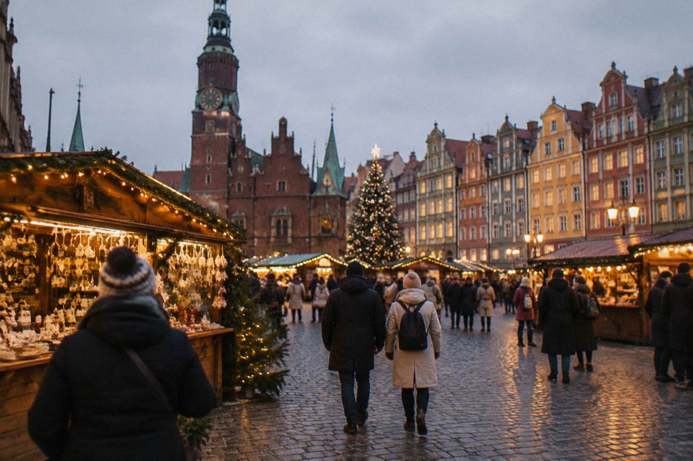 Festive wooden market stalls with glowing lights standing on a large cobblestone square in front of illuminated historic townhouses at dusk