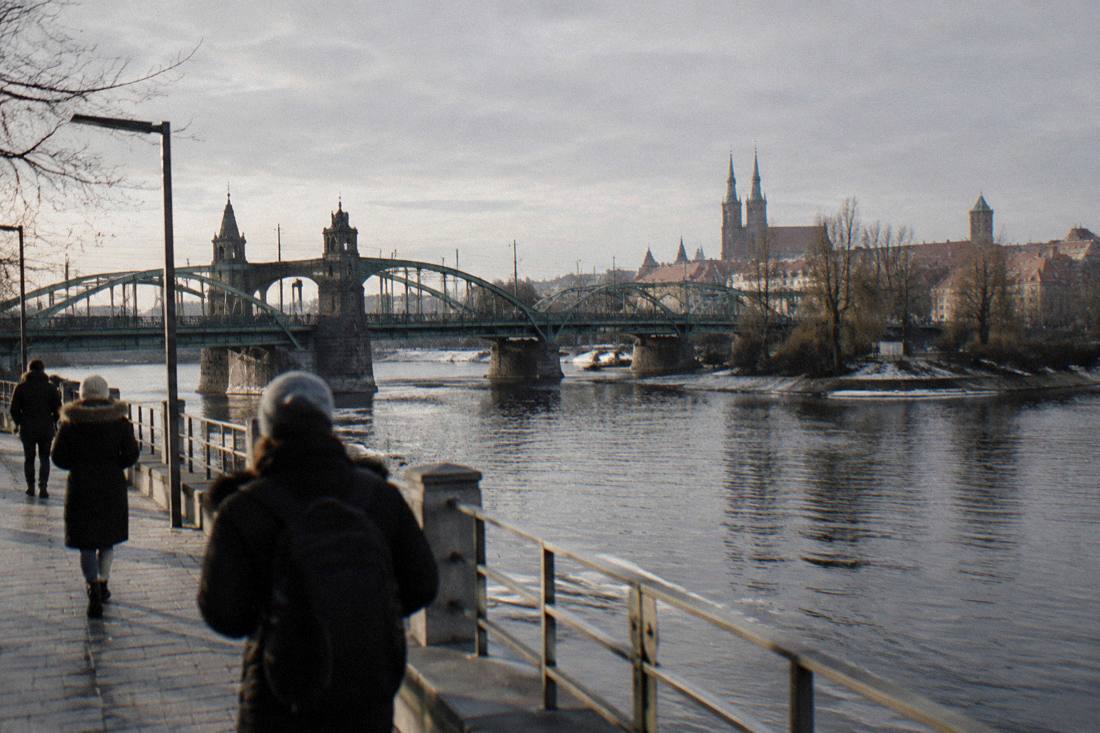 A river embankment with a historic bridge and riverside buildings under a gray winter sky