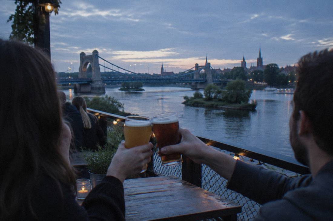 Two friends clinking craft beer glasses on a terrace beside the Odra river