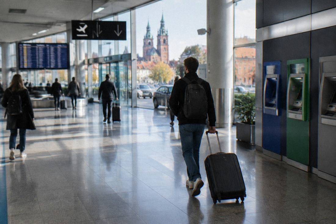 An airport arrivals hall with a traveler pulling a suitcase past bank ATM machines