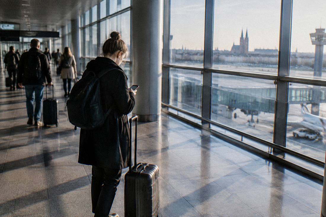 A traveler with a rolling suitcase checking a phone in a modern airport terminal corridor
