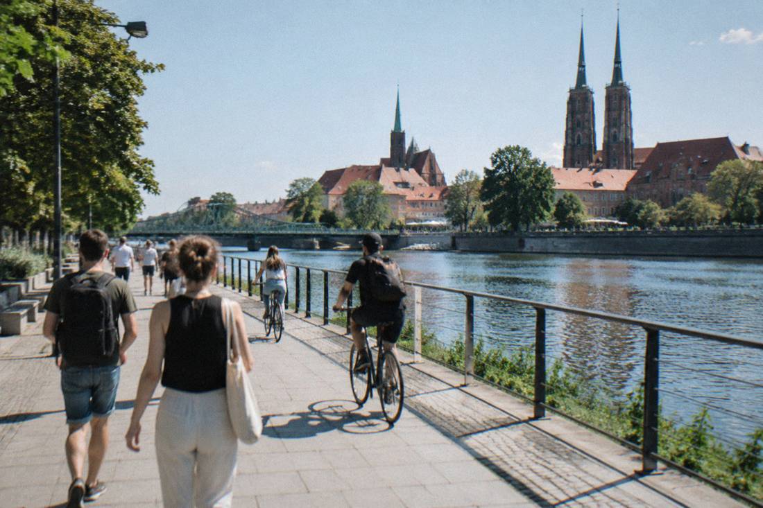 A sunny riverside promenade with people walking and cycling beside a wide river