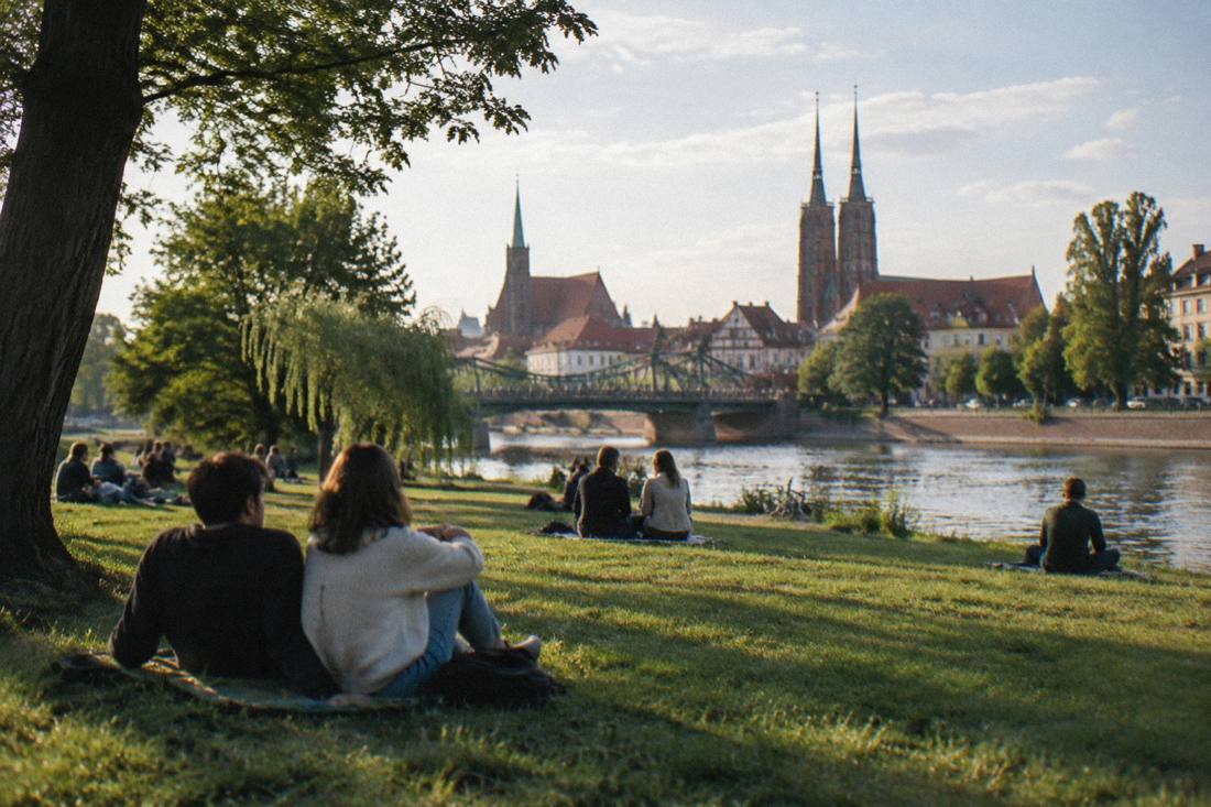 People relaxing on a grassy river island with trees and a city skyline behind