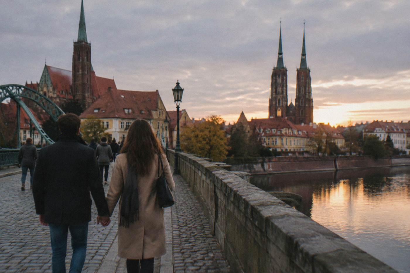 A couple walking hand in hand across an old stone bridge toward church spires at dusk