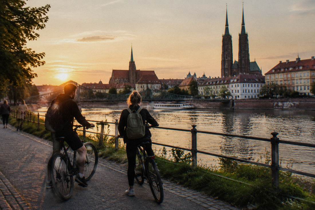 Two cyclists pausing on a riverside path with church towers behind them