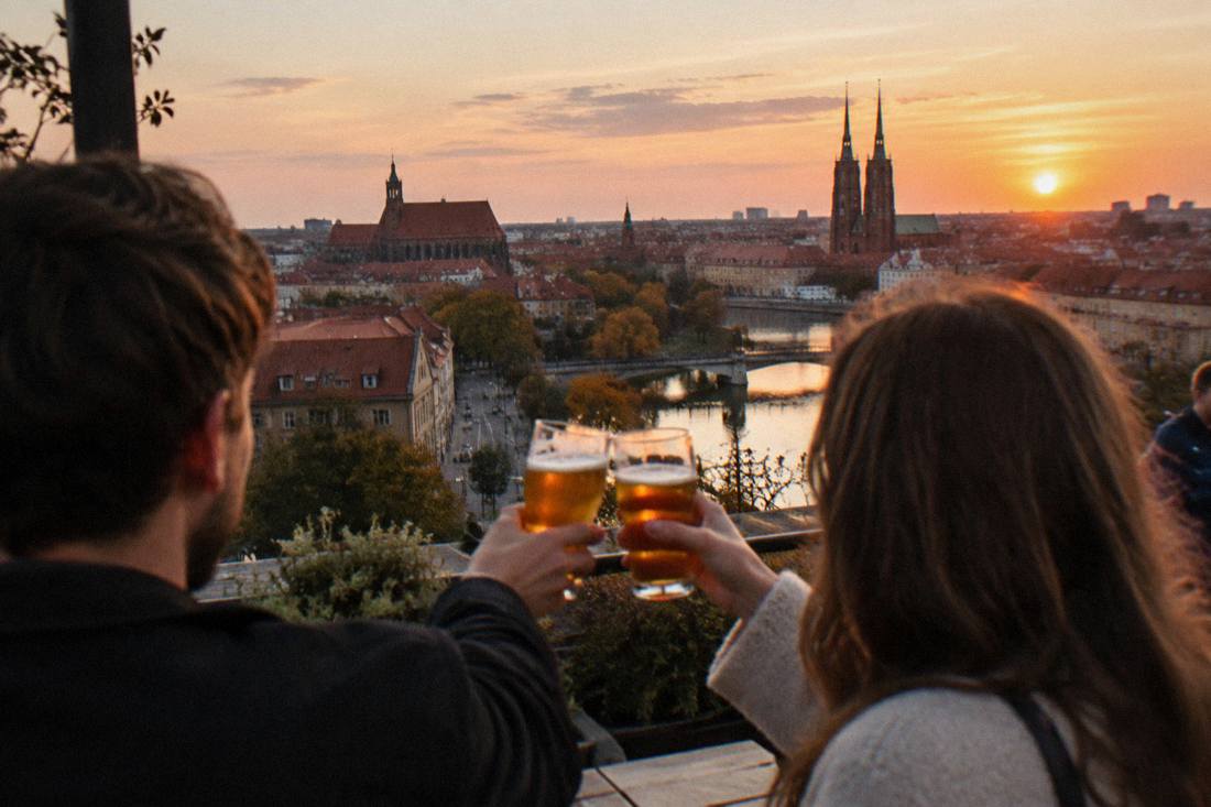 Two people toasting with beer glasses on a rooftop terrace above old rooftops at sunset
