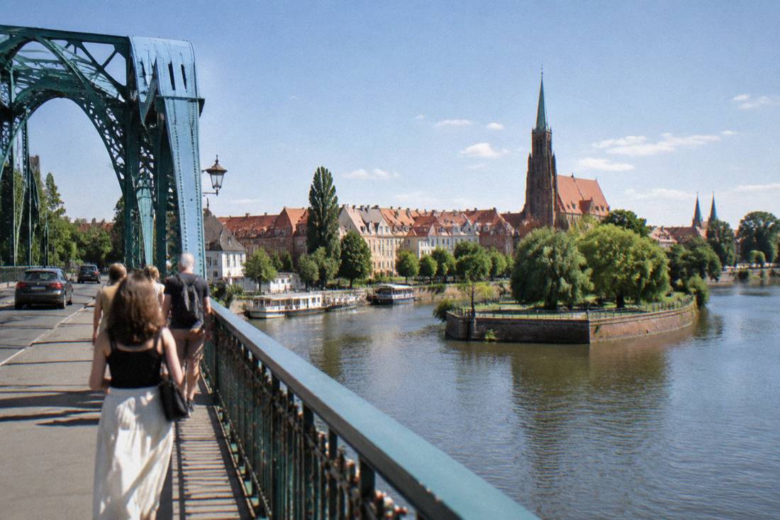 A historic bridge over the Odra river in Wroclaw with riverside townhouses and a cathedral tower in the distance