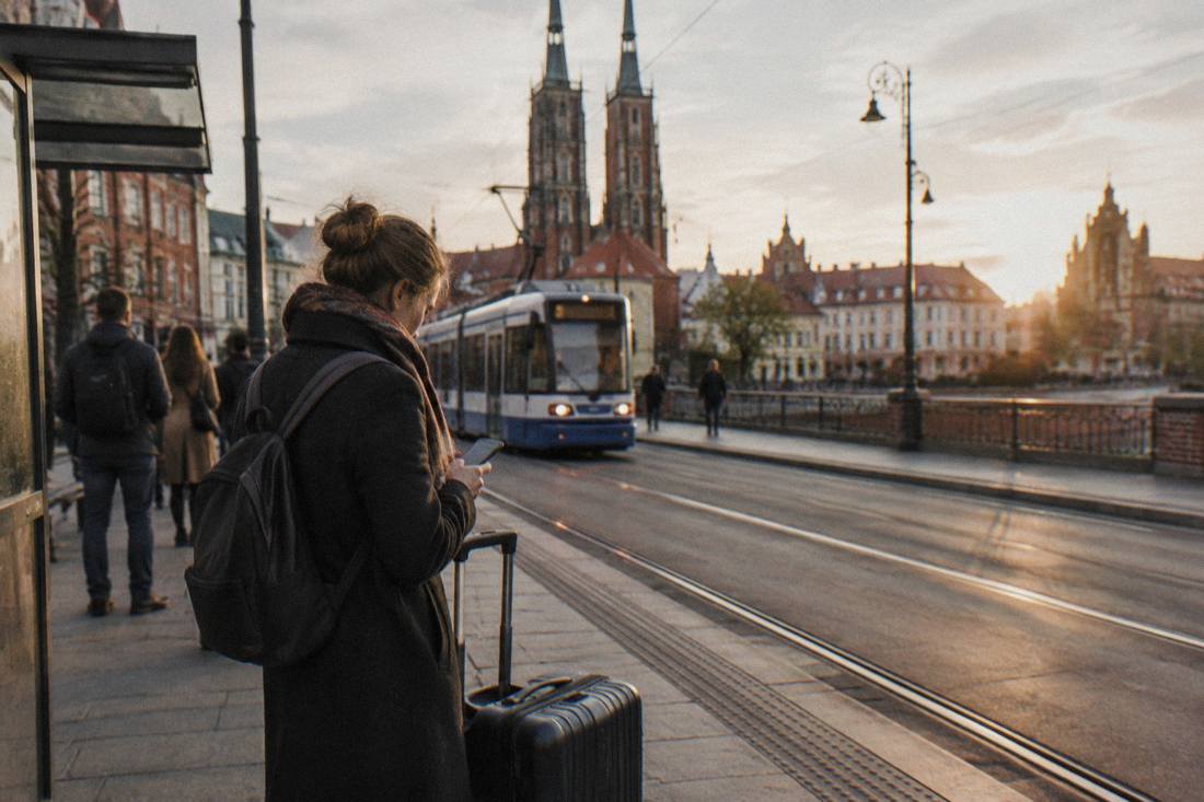 A traveler checking a phone beside a suitcase near a tram stop in a European city street