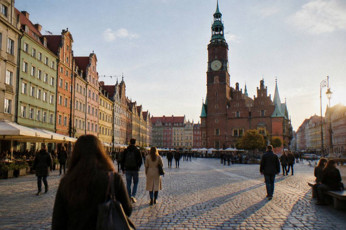 A wide cobblestone market square with colorful townhouses and a Gothic hall tower in late afternoon light